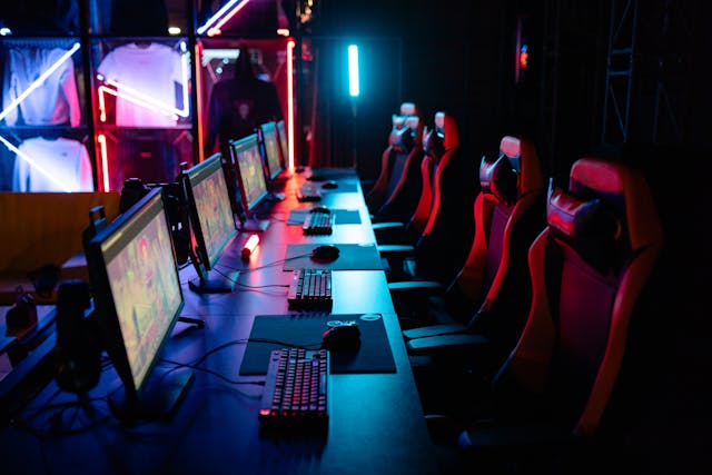 Rader med datorer och gamingstolar i ett mörkt rum med färgglad belysning, redo för e-sport.