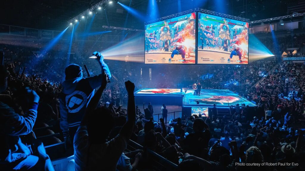 A vibrant esports event with a cheering crowd, multiple large screens displaying a fighting game, and dramatic stage lighting.