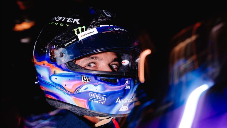 Lando Norris wearing blue Quadrant-themed helmet on a dark background