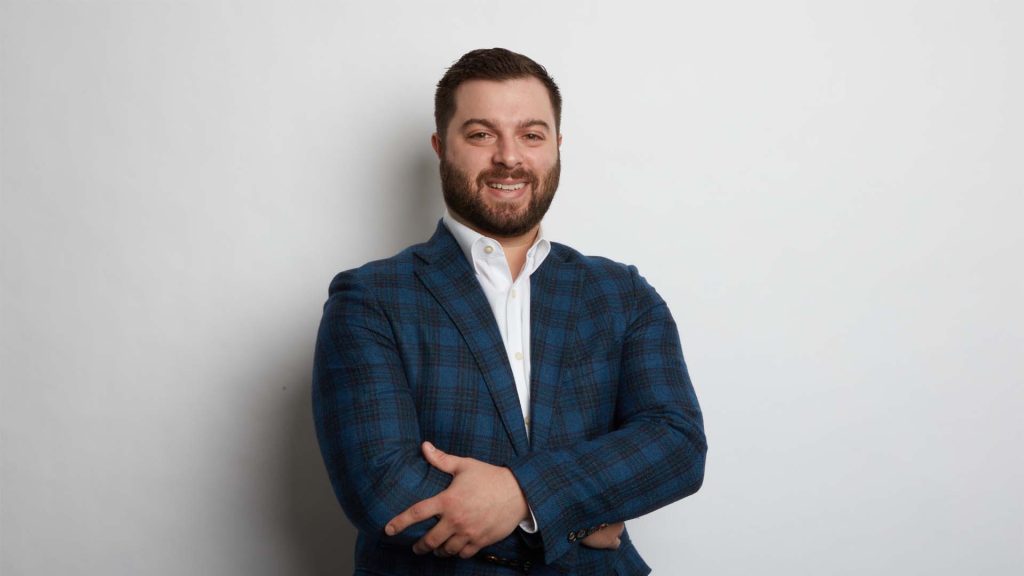 Chad De Luca wearing a blue and black checkered jacket while crossing his arms. He is standing in front of a grey wall.