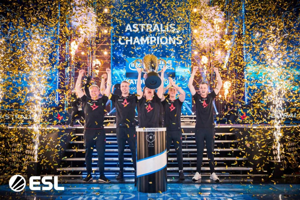 A gaming team celebrates victory, raising a trophy amidst a cascade of gold confetti and vibrant stage lights.