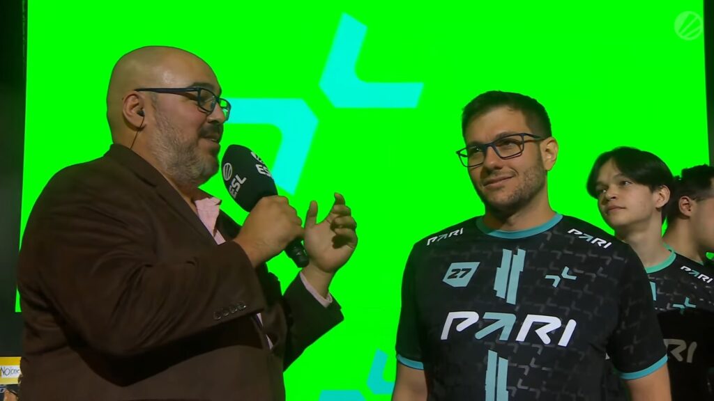 A man speaks into a microphone while standing alongside esports players wearing PARI jerseys, against a bright green background.
