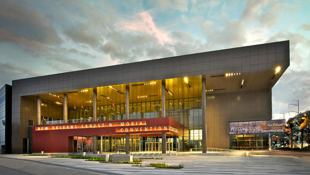 Modern exterior of the New Orleans Ernest N. Morial Convention Center, featuring large glass windows and welcoming signage, under a cloudy sky.