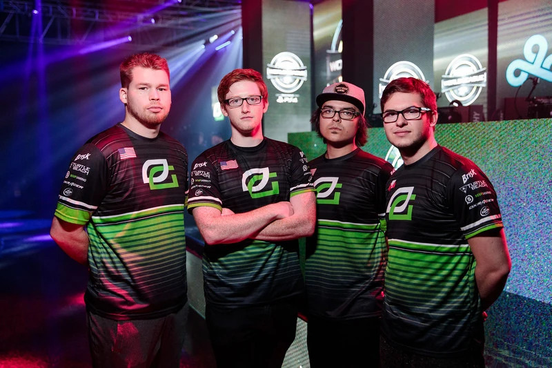 Four esports players in matching green and black jerseys pose confidently on stage, with colorful lighting and digital displays in the background.