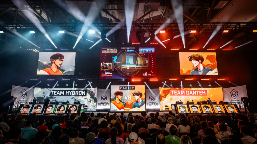 A vibrant esports arena showcasing a match between Team Hydron and Team Dantéh, with dramatic lighting and large screens displaying gameplay.