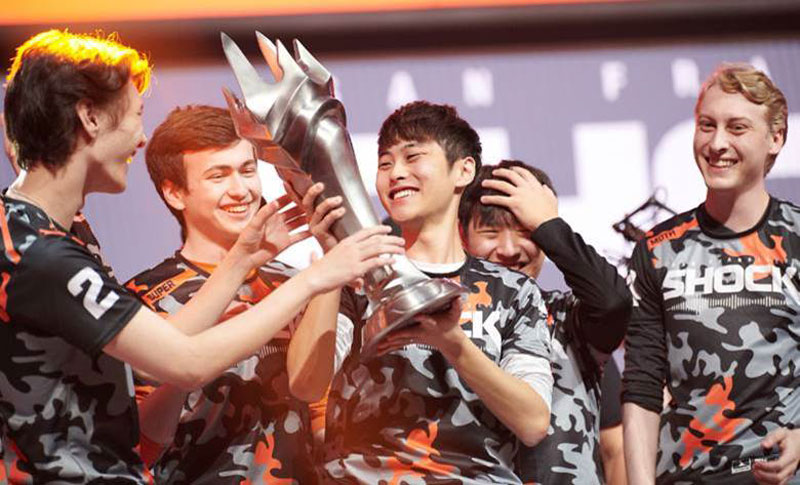 A group of esports players joyfully celebrate, holding a large trophy aloft on stage.