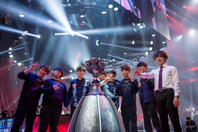 A victorious esports team stands around a large trophy on stage, with dynamic lighting and a cheering crowd in the background.