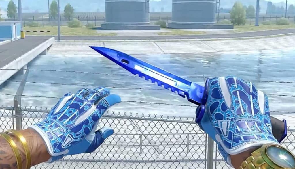 A close-up of hands with blue gloves holding a blue knife, with a blurred industrial background and a chain-link fence.