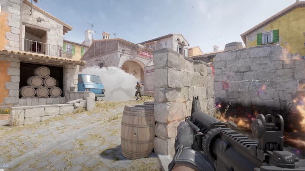 First-person view of a Counter-Strike 2 player aiming at an opponent near a stone wall and barrels on the map Inferno, with smoke and fire effects in the scene.