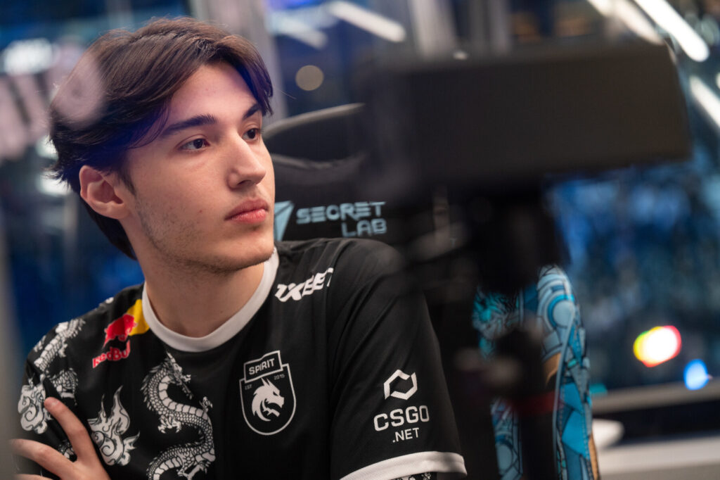 A gamer in a black team jersey, featuring a dragon design, sits in a gaming chair with a focused expression, surrounded by tech equipment.