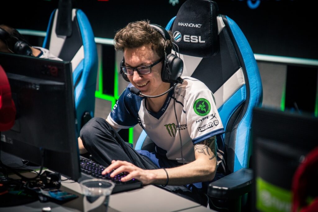 A focused gamer in a headset sits with one leg up on the desk, playing on a computer with a gaming keyboard and mouse.