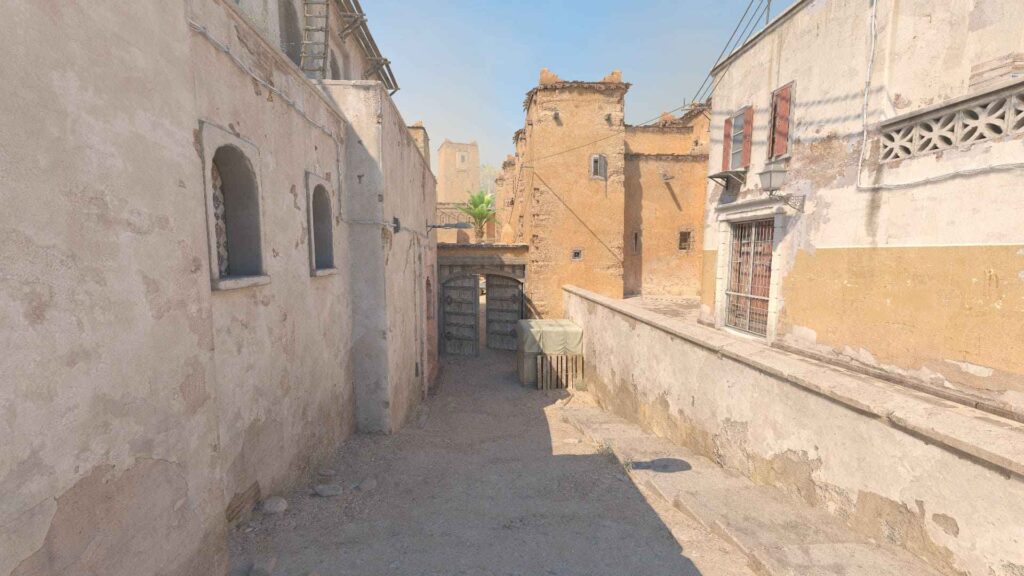 Narrow, sunlit alleyway flanked by weathered walls and arches, leading to a wooden gate with a partially visible palm tree beyond.