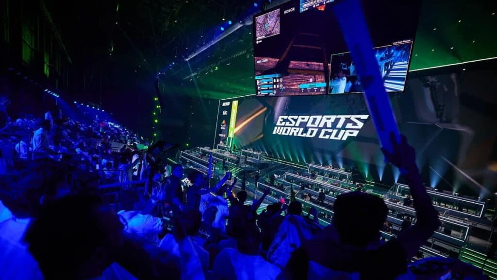 Crowd cheering at the Esports World Cup with players competing on stage and gameplay shown on large overhead screens.