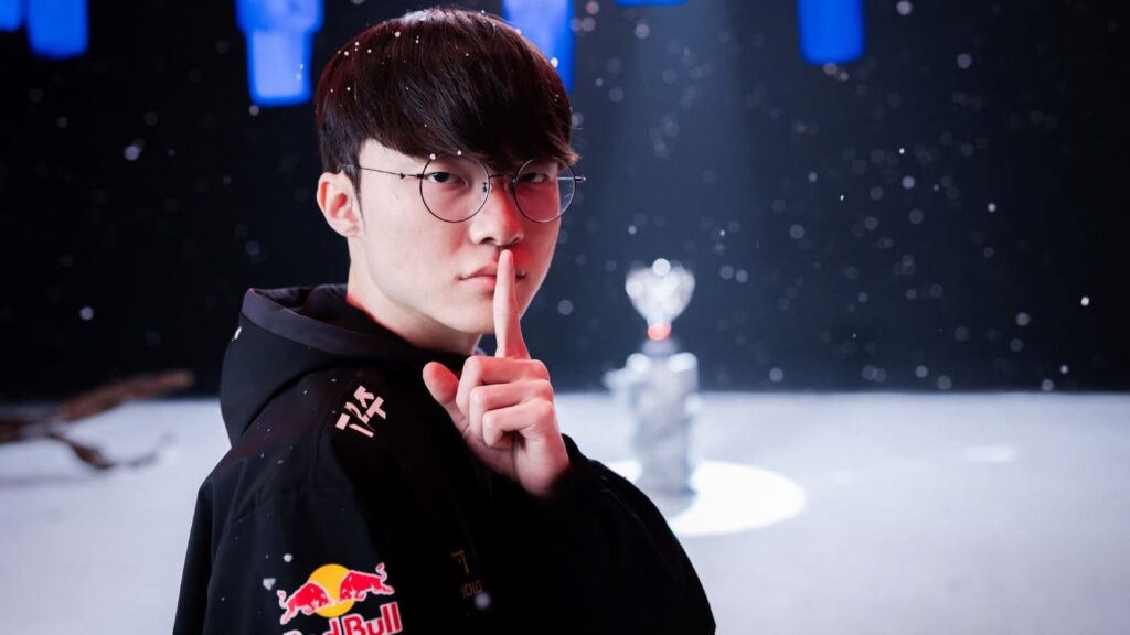 Faker in a black hoodie with a Red Bull logo poses with their finger to their lips, amidst a sparkling, snowy background and a trophy.