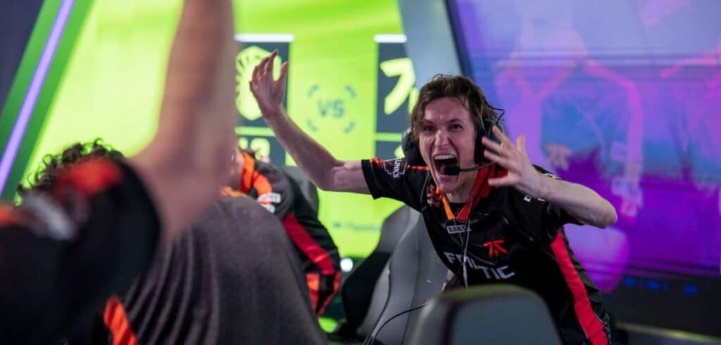 An excited esports player in a Fnatic jersey celebrates a victory, with teammates cheering and colorful game visuals in the background.