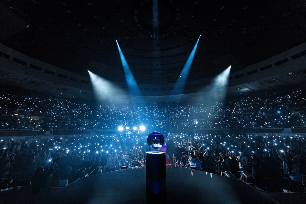 Trophy on a stage in front of a cheering crowd with lights and spotlights during an esports event in a large arena.