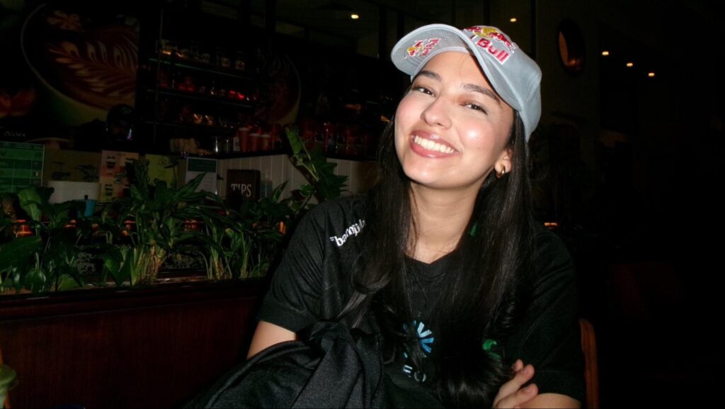 Mary from Falcons Vega wearing a cap and a black t-shirt is seated in a café surrounded by indoor plants and dim lighting.