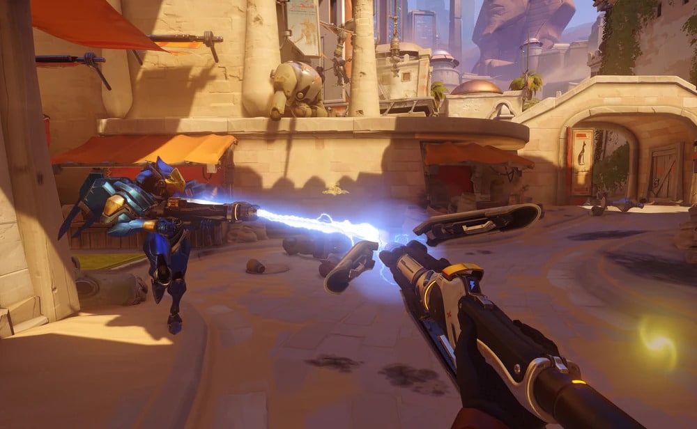 First-person view of Mercy in Overwatch 2 using her staff to damage boost Pharah in a desert-themed map.