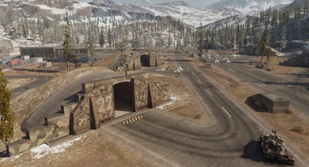 A military airfield surrounded by snow-capped mountains, featuring camouflage hangars, parked aircraft, and a tank on the road.