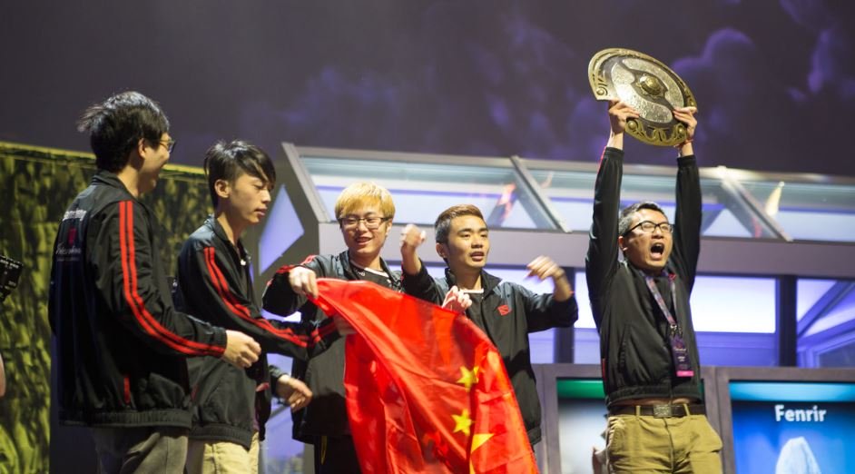 Team Newbee celebrating their victory at The International 2014, holding the Aegis of Champions trophy and the Chinese national flag on stage.