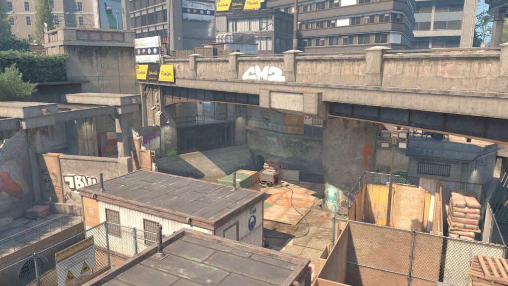 Urban industrial scene featuring concrete structures, graffiti, and fenced storage areas, under a bustling overpass in a city setting.