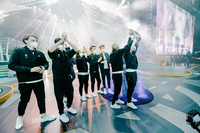 Team Spirit celebrating their victory at The International 2021 (TI10), holding the Aegis of Champions on stage amid lights and confetti, with players wearing matching tracksuits and face masks.