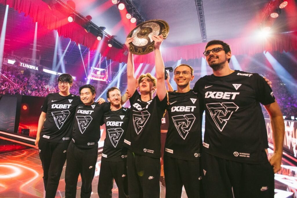 Tundra Esports celebrating their victory at The International 2022 (TI11), with players standing on stage and one team member lifting the Aegis of Champions trophy, under bright arena lights and cheering crowd.