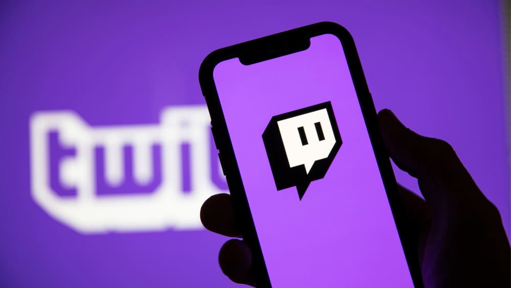 A hand holds a smartphone displaying the Twitch logo, with the iconic purple background and the Twitch name in the backdrop.