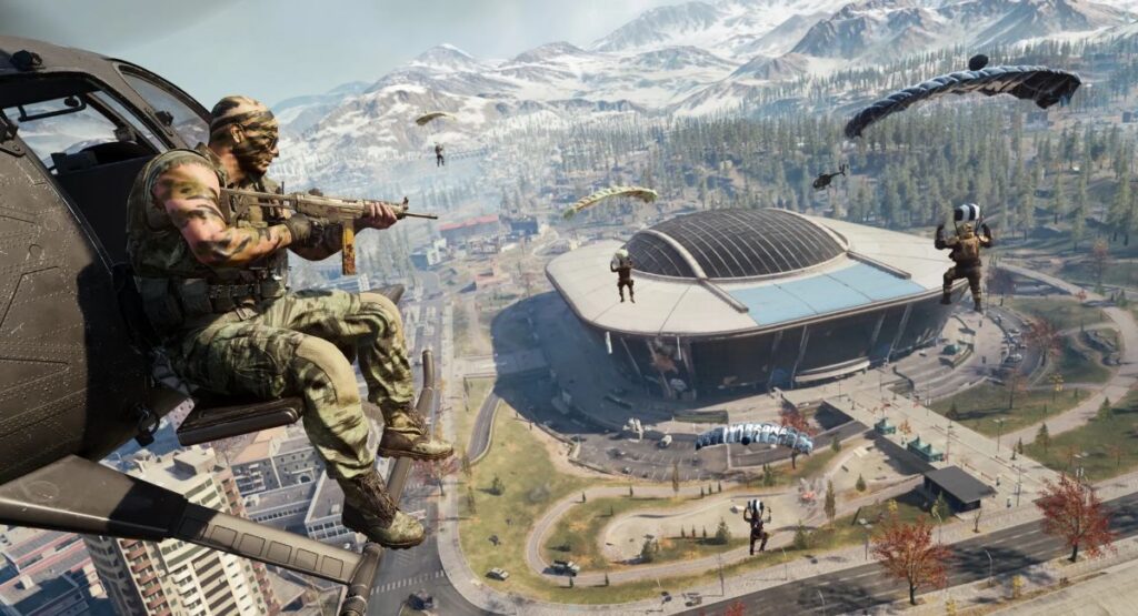 A soldier hangs from a helicopter, armed with a rifle, while several others parachute towards a large stadium below, set in a mountainous landscape.