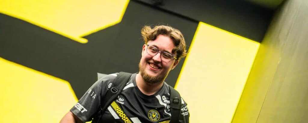 ZywOo from Team Vitality smiling in front of a large yellow and black backdrop, wearing his team jersey and backpack.