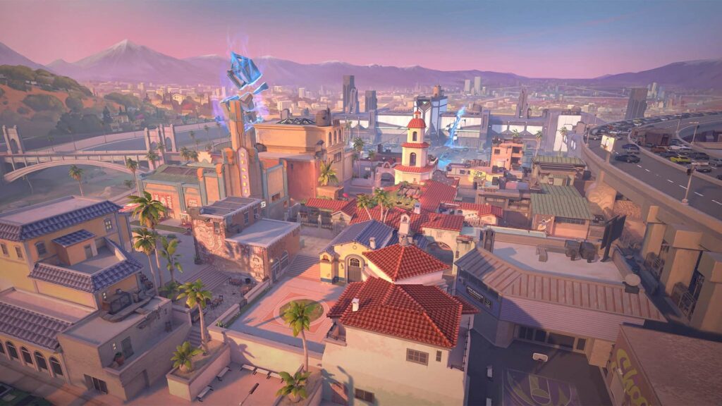 A vibrant urban landscape of a Sunset map of VALORANT with colorful buildings, palm trees, and a distant mountain range, set against a sunset sky.