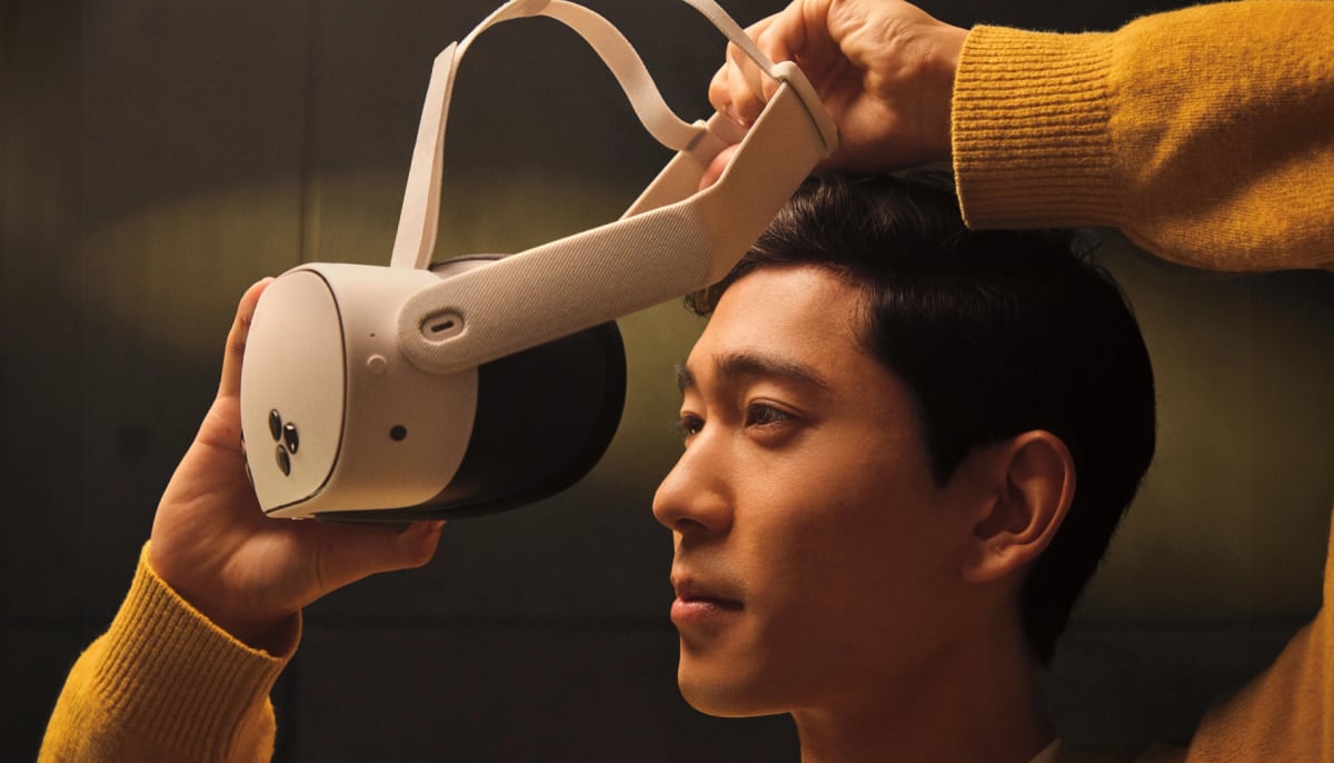 A person in a yellow sweater lifts a virtual reality headset towards their face, focused and curious