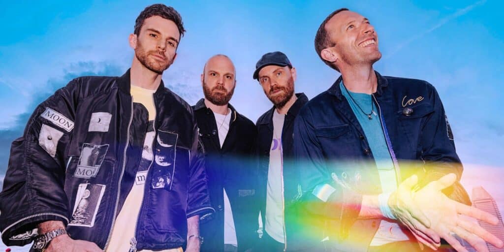 Four members of Coldplay stand against a vibrant sky with a rainbow glow across the image