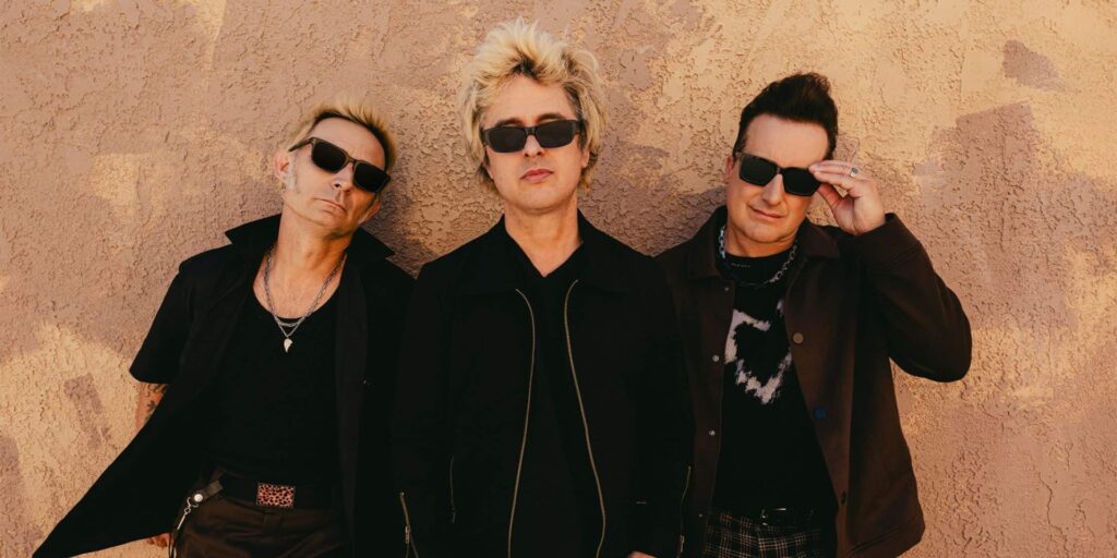 Green Day's members wearing sunglasses stand against a textured beige wall. They exude a cool, casual vibe, with relaxed postures and neutral expressions