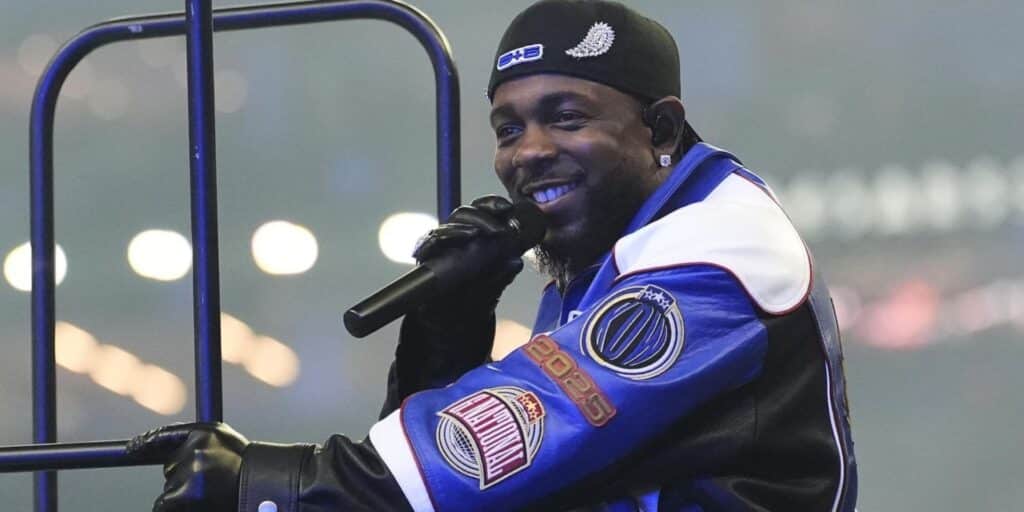 Kendrick Lamar in a blue jacket with patches and a black beanie smiles while holding a microphone
