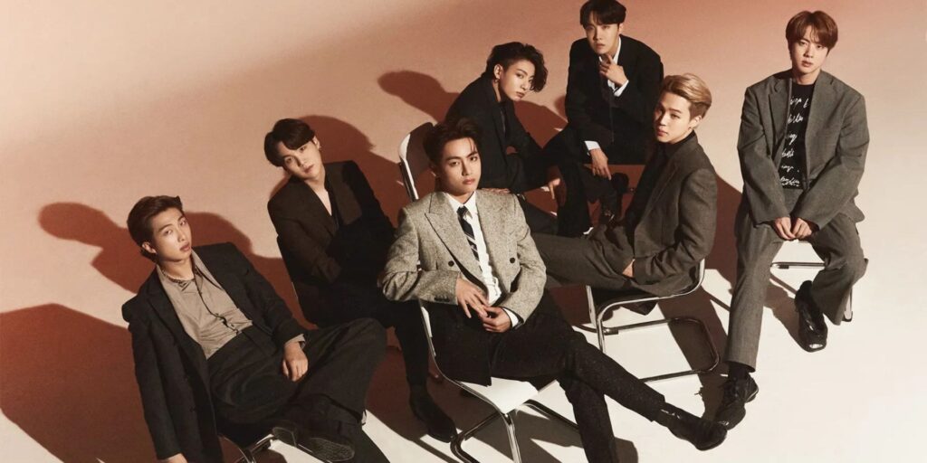 BTS in stylish suits are seated in a semi-circle on chairs, casting long shadows on the floor