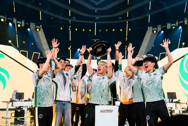 A joyous esports team celebrates on stage, wearing matching jerseys. One member raises a trophy high, with dynamic stage lights and monitors in the background