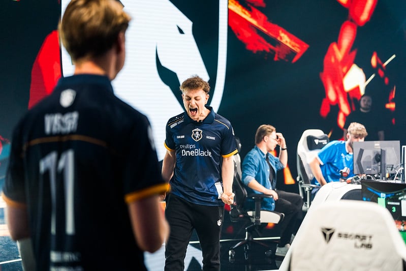 An esports player in a navy jersey celebrates passionately on stage, clenching his fist