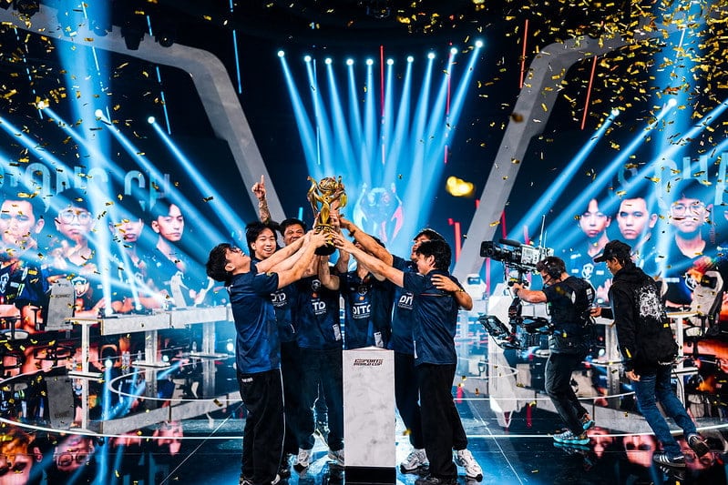 A team of five people in blue jerseys joyfully lift a trophy on stage, surrounded by confetti and vibrant stage lights