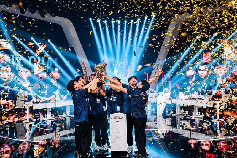 A team of esports players in blue jerseys celebrate under a shower of golden confetti, raising a trophy, on a brightly lit stage with a joyful atmosphere