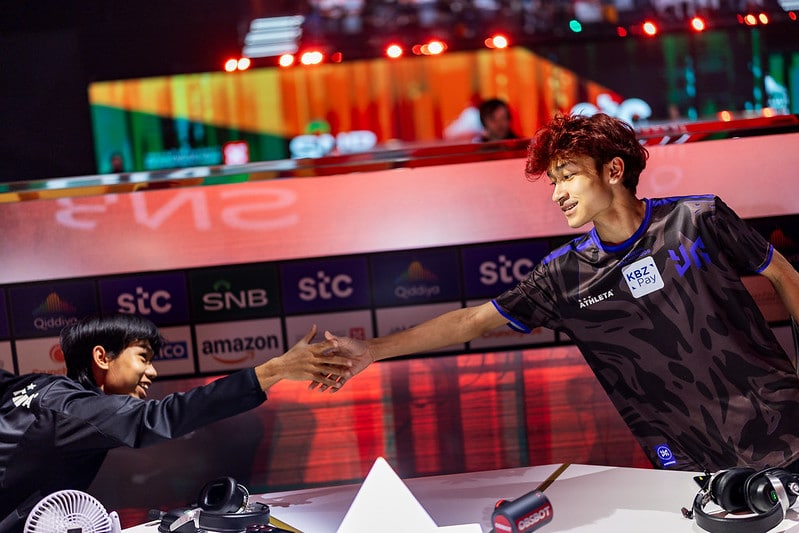 Two esports players exchange a friendly handshake across a gaming table, surrounded by branded backgrounds and equipment, conveying sportsmanship