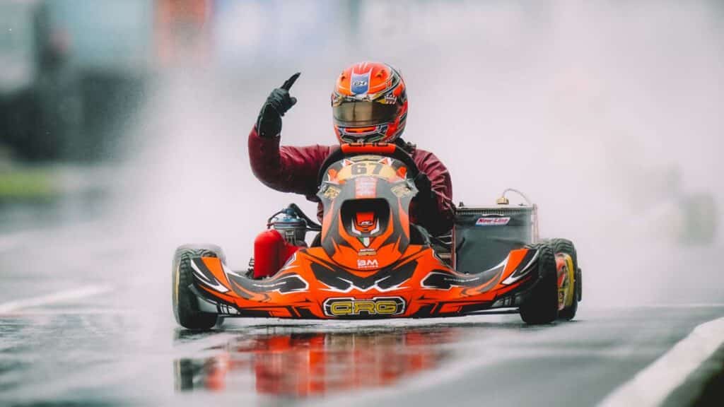 Image of go kart driver pointing finger in the air while driving a go kart. Spray from a wet track is being dispersed in the background