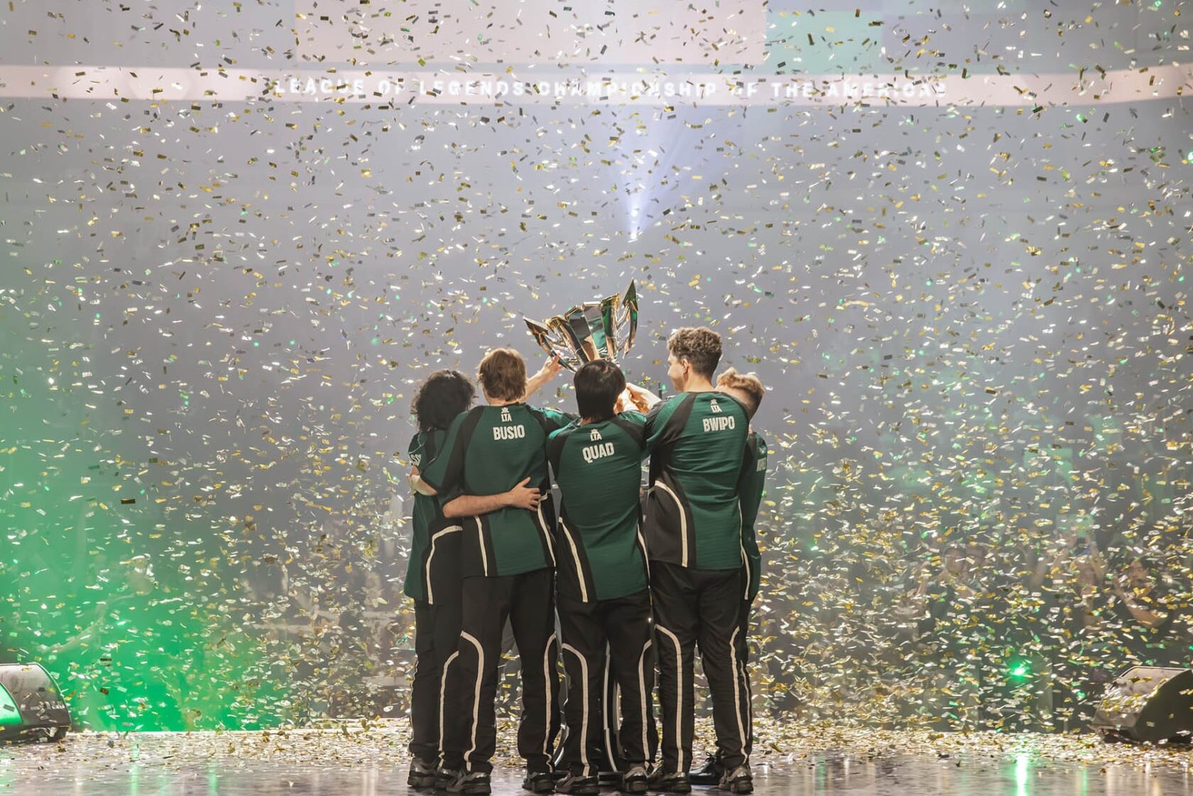 FlyQuest team lifting the LTA North trophy on stage with confetti and crowd in the background.