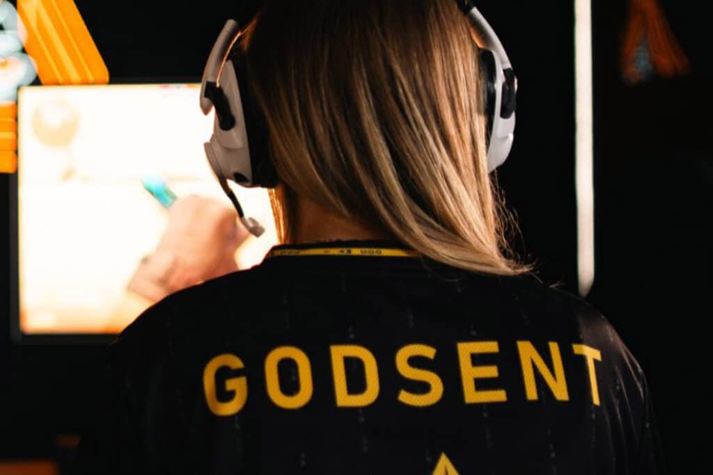 Image of blonde-haired person wearing a black jersey with GODSENT written on the back. A blurred gaming monitor is in the background