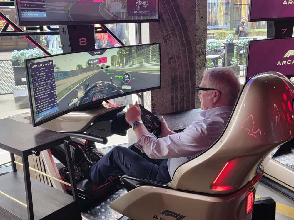 Richard Parker con una camicia bianca e gli occhiali da sole gioca un gioco di simulazione delle corse di Formula 1 in un ambiente arcade