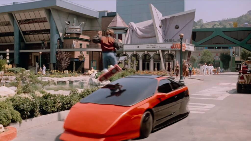 La scena del film Ritorno al futuro mostra Marty McFly su un hoverboard che sorvola un'auto futuristica rossa e nera in una strada cittadina vivace e movimentata.