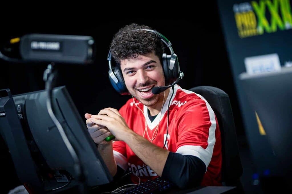 A smiling man in a red esports jersey sits at a gaming desk with a headset and microphone