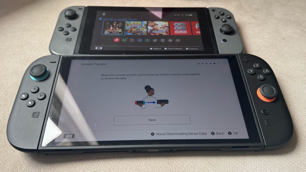 L'immagine mostra il processo di trasferimento dei dati tra due console Nintendo. A giudicare dall'interfaccia e dalla levetta arancione sulla console inferiore, questo è il modello originale di Nintendo Switch (probabilmente la versione V2 o OLED) e il nuovo modello di Nintendo Switch 2.