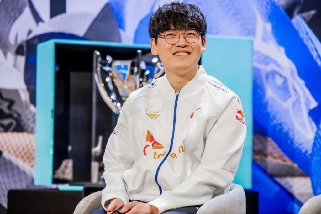 Gumayusi mit Brille und weiß-blauer E-Sport-Jacke mit Sponsorenlogos (Samsung Odyssey, Red Bull, T1) sitzt lächelnd vor einer großen silbernen Trophäe