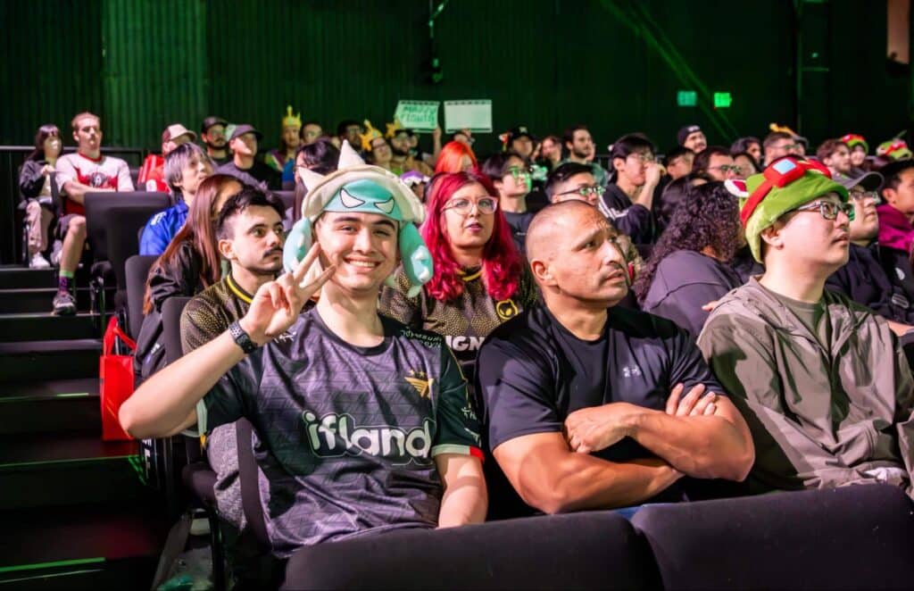 Folla di entusiasti fan degli eSport in un'arena poco illuminata, vestiti con costumi e cappelli colorati come Pokémon che esultano con segni e segni di pace, seduti in file sfalsate sotto l'illuminazione verde.